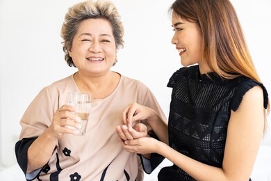 caregiver woman helping old elderly woman taking medicine pill with water on the bed and check up her at home.