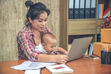 pretty young single mom working at home on a laptop computer while holding her baby girl sitting on her lap enjoying playing computer.