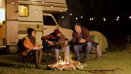 man singing a song on guitar for his friends around camp fire in a cold night of autumn in the mountains. retro camper van. light bulbs.
