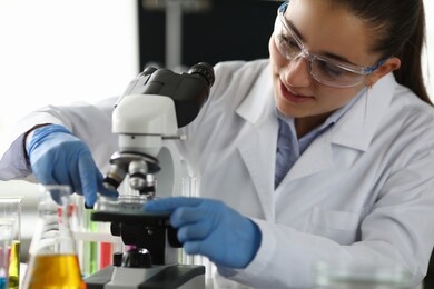 portrait of research technician putting some substance under microscope in laboratory office. smiling woman in protective uniform. science and investigation concept
