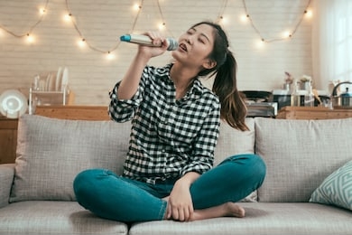 young asian girl singing with microphone on sofa at home kitchen. beautiful lady love music enjoy karaoke relax in couch in apartment. chinese female having fun alone in modern house interior.