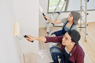 professional young asian wall painters in denim aprons using foam rollers when applying white paint