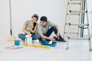 excited pretty young vietnamese woman opening can of wall paint and showing color to boyfriend