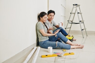 young asian couple sitting at wall, drinking coffee and discussing plan of apartment renovation on tablet computer