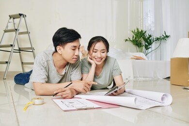 happy young vietnamese couple with blueprint and tablet computer lying on floor in their new house and discussing design ideas