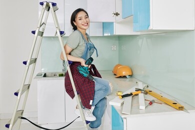 smiling attractive young vietnamese woman sitting on ladder with screwdriver in hand in kitchen where she is assembling cupboards