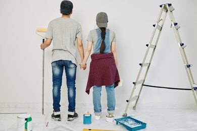 young couple holding hands and looking at wall they are going to paint, view from the back