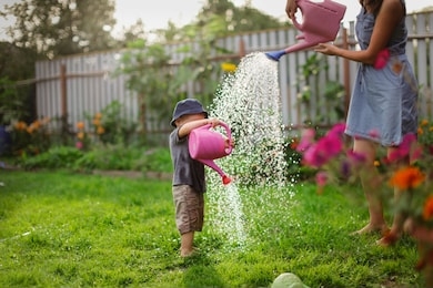 mom and boy child water the garden together, mom’s little gardener assistant, taking care of children and garden. mother watering her son from watering can, take care of trees and plants, wet child