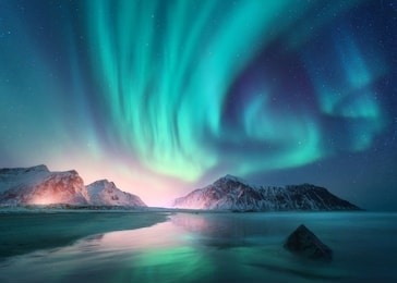 aurora borealis over the sea, snowy mountains and city lights at night. northern lights in lofoten islands, norway. starry sky with polar lights. winter landscape with aurora, reflection, sandy beach 