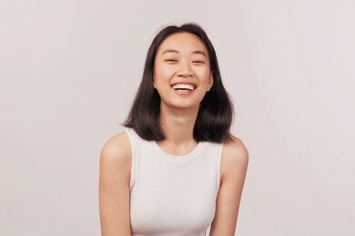 girl laughs at something funny closes eyes with pleasure. businesslike young woman asian appearance with black hair and brown eyes dressed in short shirt stands isolated white background in studio.