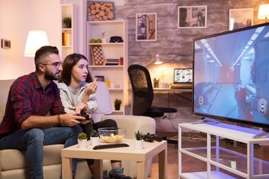boyfriend playing video games on television using controller while girlfriend is eating chips. couple sitting on sofa.