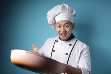 portrait of asian male chef smiling happy while holding kitchen tools, chef holding spatula and pan, ready to cook, against blue background