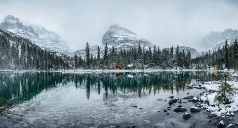 panorama of wooden lodge in pine forest with heavy snow reflection on lake o'hara at yoho national park, canada