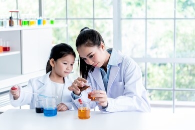 the pretty asian teacher is teaching a 6 year old girl, about scientific experiments on white table in a science lab, with blur background to children and education concept.