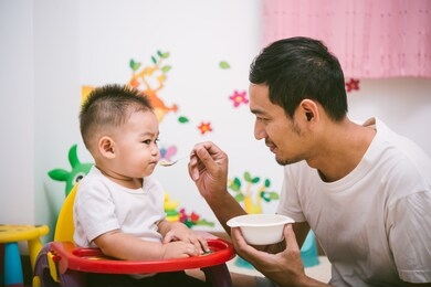 father acting mom feeding his son baby 1 year old on chair in the house