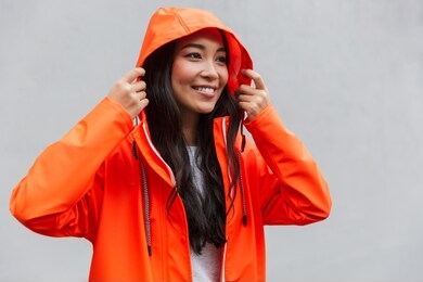 smiling young asian woman wearing raincoat walking outdoors in the rain, posing in a hood