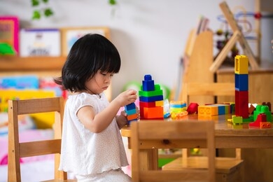 asian adorable laughing little child girl playing a colorful plastic block. sitting on a wooden chair. in a room at home or kindergarten school. baby age 2-3 years old.
