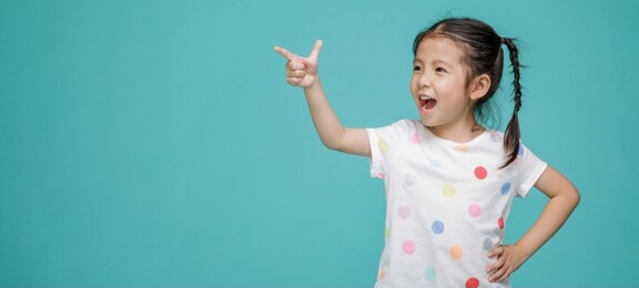 beautiful smiling asian little girl pointing hand to blank speech bubble, empty space in studio shot isolated on colorful blue background, educational concept for school