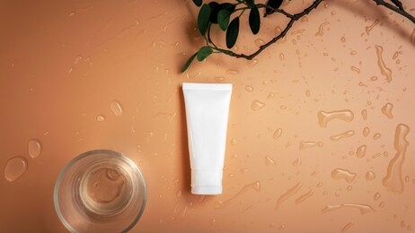 top view empty organic facial skincare white tube product next to shining glass cup with branch and leaves on droplets orange brown background.