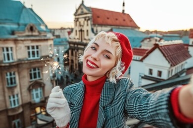 happy smiling girl with sparkler takes selfie on roof top with beautiful view of old european city. winter holidays, christmas vacation, new year, travel, tourism concept. copy, empty space for text