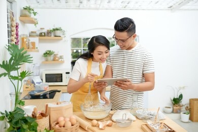 young asian man and woman together cooking cake and bread with egg, looking menu from tablet in the flour happy relaxing in at home