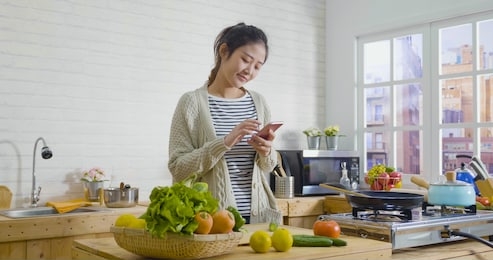 picture of beautiful asian chinese woman cooking vegetables in cozy wooden kitchen while looking recipe on smartphone. young girl browsing website on tutor cooking class and smiling checking fruits.