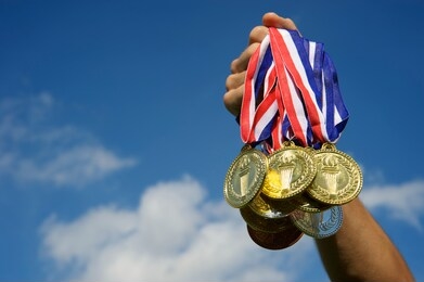 hand of an unrecognizable athlete holding up a bunch of gold, silver, and bronze medals in bright blue sky