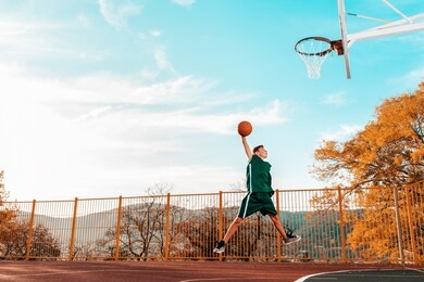 sports and basketball. a young teenager in a green tracksuit jumps and throws a ball into the basket. blue sky and court in the background. copy space