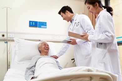 two young friendly asian doctors talking to senior asian male patient at bedside in hospital