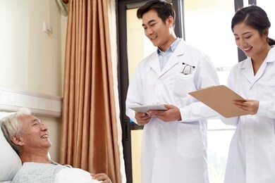 two young friendly asian doctors talking to senior asian male patient at bedside in hospital