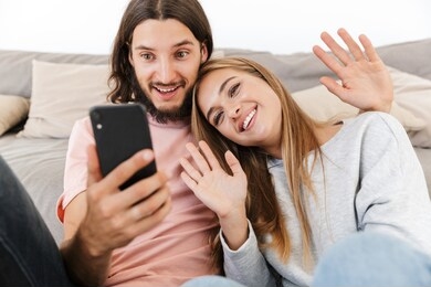 happy lovely couple relaxing on a couch at home, having a video call while holding mobile phone, waving hands