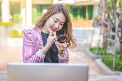 portrait of beautiful asian woman putting on her foundation while looking in the mirror within the makeup compartment, with laptop computer in front, wearing a pink and white blazer and black shirt