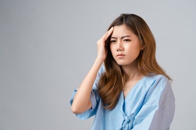 portrait of a beautiful asian woman in patient gown raised her right hand and touch her head feeling like headache and fever sickness on gray background with copy space, concept healthcare insurance.