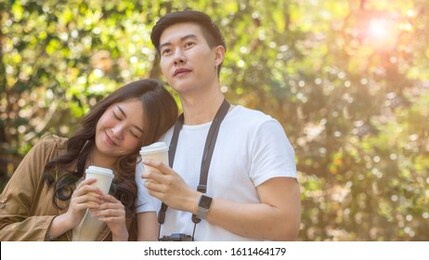 young asian couple, newly weds or lovers having hot coffee in the paper cup while walking and enjoy the romantic time together in the park, forest or camping sight. love, togetherness and valentine's
