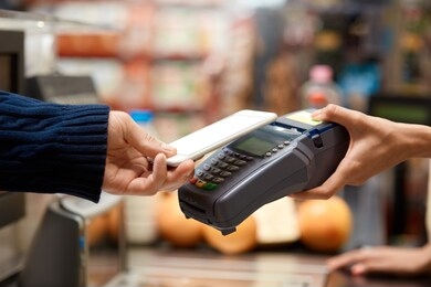 cropped view of young adult man standing on checkout, paying for shopping in grocery store, using modern smartphone while cashier holding terminal in hand