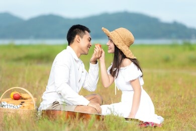 lifestyle couple happy honeymoon in sunny time. asian young couple sitting picnic and relax in the meadow and field near river and mountain in holiday.  romantic and in love.   lifestyle concept