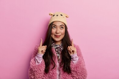 look up. attractive brunette woman points index fingers upwards, demontrates cool item with pleasant tender smile, wears pink coat, stands over rosy pastel background, likes new promo or advert