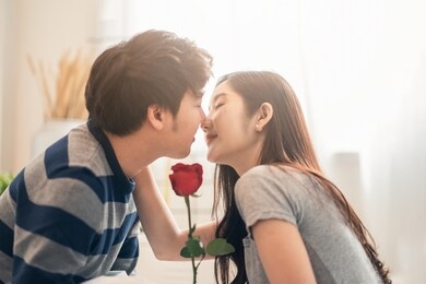 close up shot asian pretty woman and handsome man holding rose flower together sitting on bed in bedroom, closing eyes, smile and kissing with romantic moment. couple lover and lifestyle in valentine.