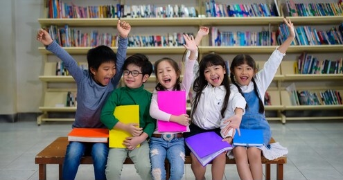 portrait a group of young little asian kid hold  colourful of book in school library and sitting on table with shelf of book, asian kid education concept