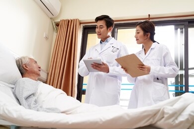 two young friendly asian doctors talking to senior asian male patient at bedside in hospital