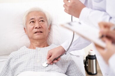 asian old man lying in bed in hospital ward being examined by doctor using stethoscope