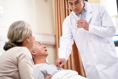 asian elderly male patient lying in bed being examined by young doctor in hospital ward