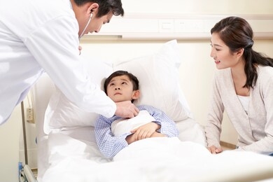 five year old asian kid lying in bed in hospital ward accompanied by mother and being examined by pediatrician using stethoscope