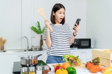 cheerful beautiful woman hand holding a smartphone and salad bowl with ladle and various green leafy vegetables on the table. talking on the mobile with a friend in the morning in the kitchen