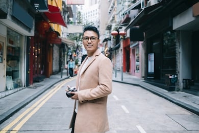 side view of glad asian male tourist in trendy beige greatcoat and stylish eyeglasses photographing with camera on street road and looking away  