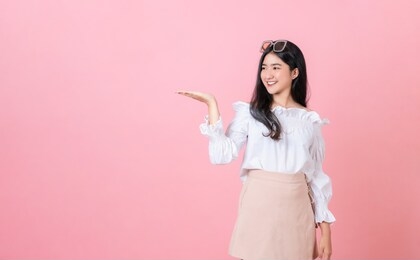young smiling asian woman stand with open palms and looking to copy space on pink background.