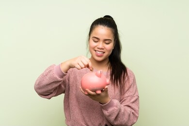 young teenager asian girl over isolated green background holding a big piggybank