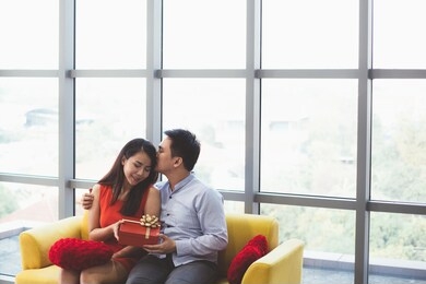 a man give present gift box to his girlfriend on sofa and kiss on her head at home, asian attractive couple have a romantic dating on valentine's day, birthday, christmas or anniversary concept
