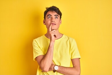 teenager boy wearing yellow t-shirt over isolated background with hand on chin thinking about question, pensive expression. smiling with thoughtful face. doubt concept.
