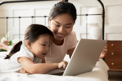 happy young asian mom and little ethnic daughter lying in bed at home watch video on modern laptop, smiling vietnamese mother or nanny relax in bedroom with small biracial girl child using computer